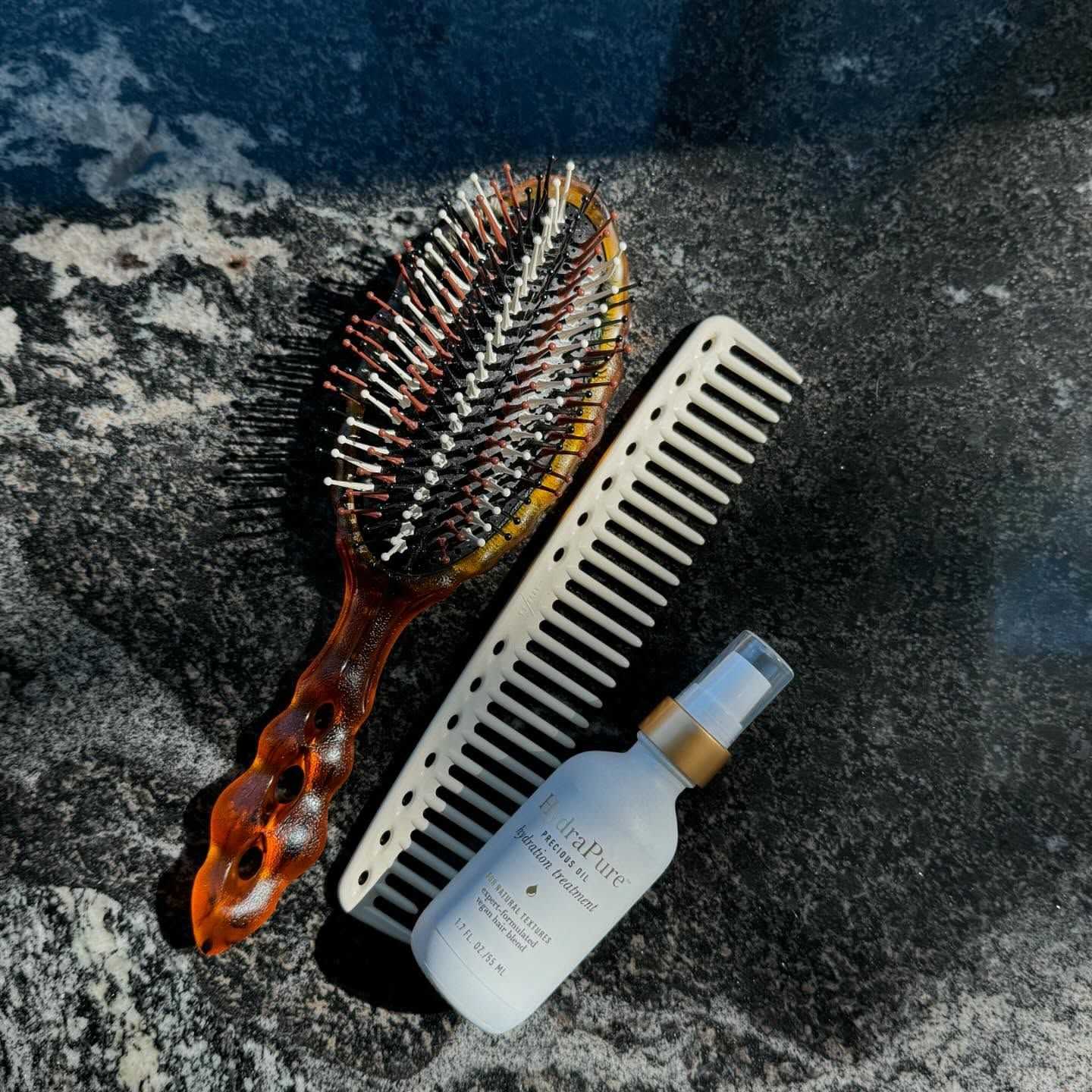 Hairbrush, comb, and bottle of hair product on a dark marble surface.