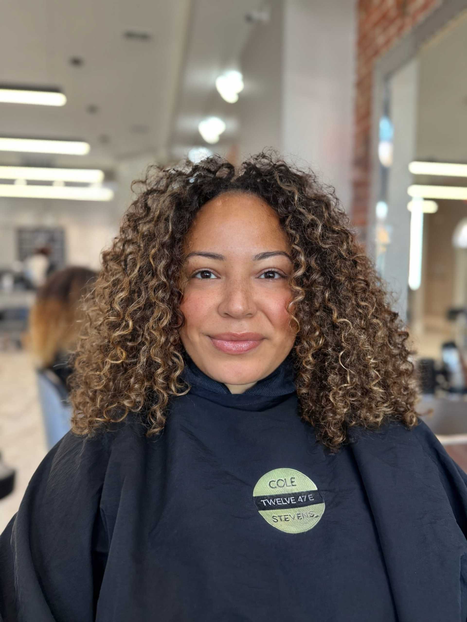 Woman with curly hair smiling in a salon, wearing a black smock.