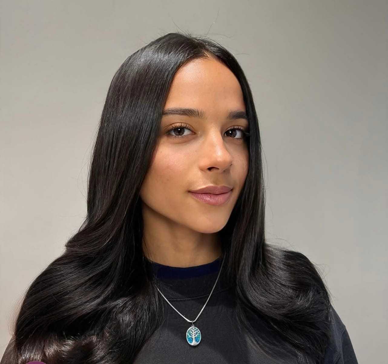 Young woman with long dark hair, wearing a necklace, against a plain background.