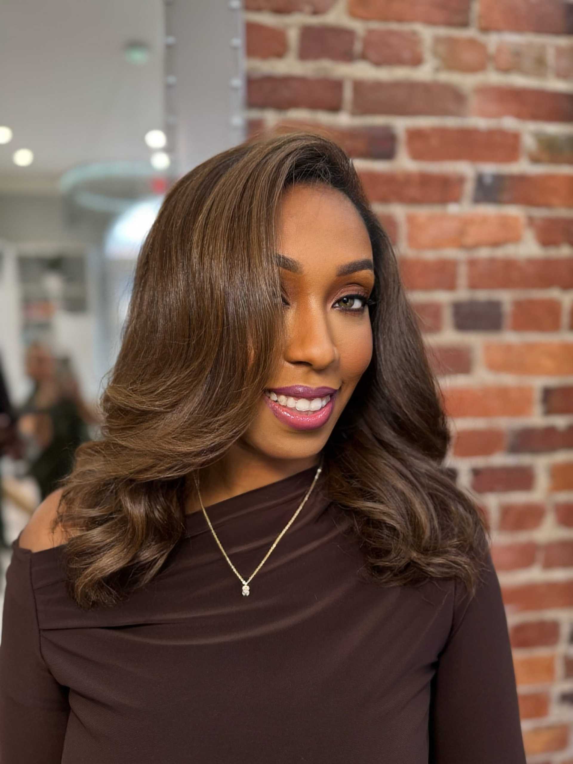 Woman in a brown dress smiling against a brick wall background.